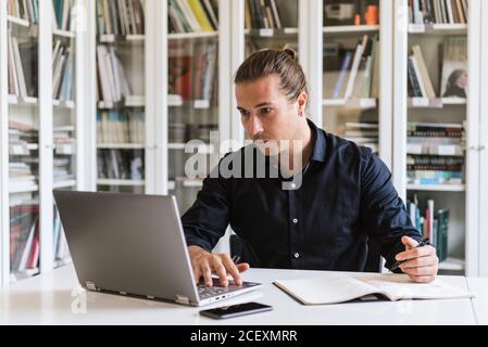 Focused businessman in formal shirt sitting at table while doing paperwork and typing on notebook in bright workspace Stock Photo