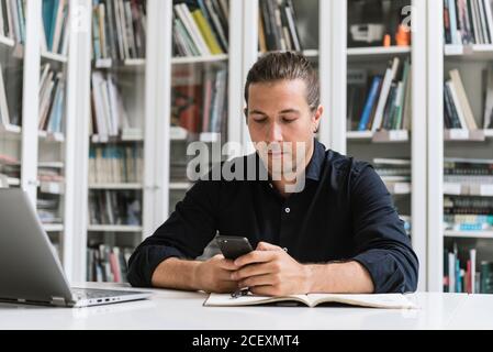 Concentrated male entrepreneur sitting at table with laptop and browsing smartphone while working on project in contemporary workplace Stock Photo
