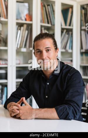 Handsome male office worker in formal wear sitting at table in modern workspace and looking at camera Stock Photo