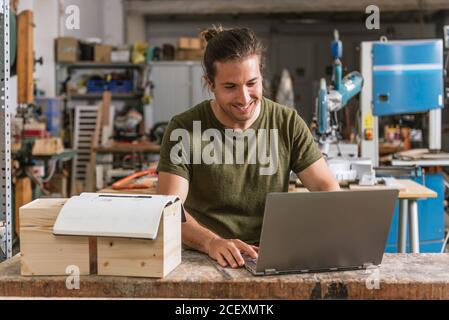 Content male carpenter sitting at workbench and working on netbook on project in modern workshop Stock Photo