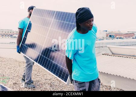 African American workers walking with solar panel on sunny day in industrial area Stock Photo