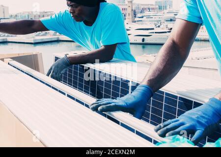 Crop unrecognizable African American male workers in protective gloves installing contemporary solar panels in industrial area of plant during daytime Stock Photo