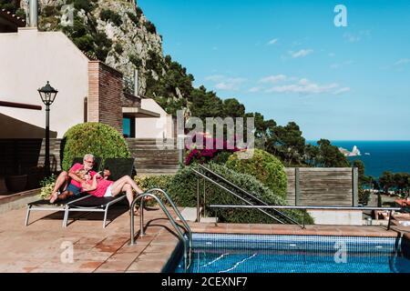 Loving senior couple of tourists relaxing on deck chairs at poolside and browsing smartphone together during summer vacation Stock Photo
