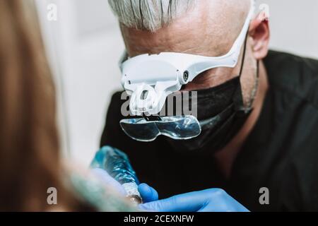 Busy male tattoo master in gloves and mask using machine with ink and creating picture on arm of client in salon Stock Photo