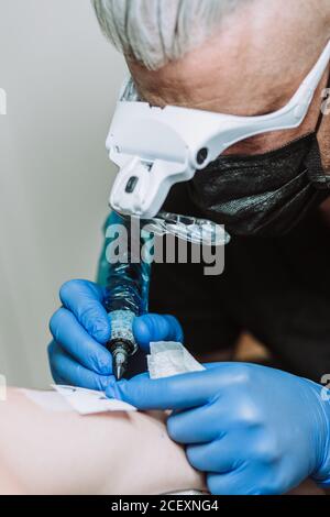 Busy male tattoo master in gloves and mask using machine with ink and creating picture on arm of client in salon Stock Photo