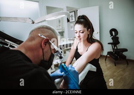 Busy male tattoo master in gloves and mask using machine with ink and creating picture on arm of client in salon Stock Photo