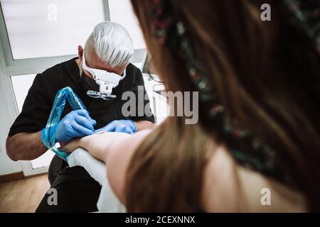 Busy male tattoo master in gloves and mask using machine with ink and creating picture on arm of client in salon Stock Photo