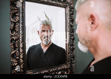Confident brutal middle aged bearded male with gray hair in black clothes  looking at reflection in framed mirror Stock Photo