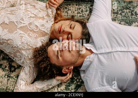 From above romantic newlywed young couple in stylish clothes lying opposite with face to face with eyes closed cuddling on vintage rug on sandy beach Stock Photo