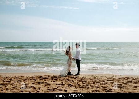 Side view of groom and bride in beautiful wedding clothes standing on sand near waving sea during romantic journey holding hands and looking at each other smiling Stock Photo
