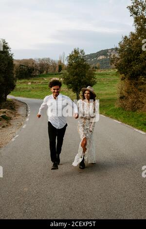 Full body delighted young romantic couple in elegant wedding clothes holding hands and running on asphalt road in green mountainous terrain in spring day in Spain Stock Photo