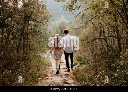 Back view of unrecognizable young bride and groom in stylish wedding outfits walking together on narrow stony path in mountainous forest in Morro de Labella in Spain Stock Photo