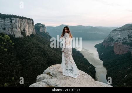 Full length back view of charming young Woman in translucent white wedding gown looking at camera while standing on edge of rocky cliff against spectacular scenery with mountains and lake of Morro de Labella in Spain Stock Photo