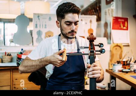 Young professional male master using peg hole reamer while repairing and tuning string instrument in workshop Stock Photo