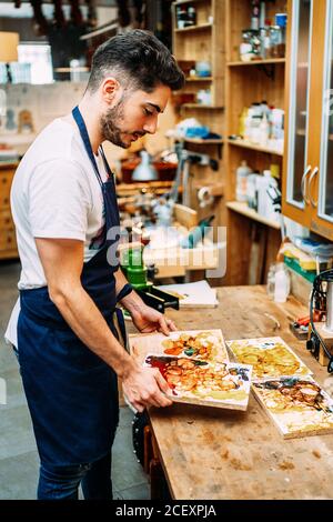 Side view of young male artisan luthier standing at counter and holding wooden boards with mixed lacquers for polishing musical instrument in workshop Stock Photo