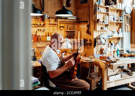 Side view of mature male luthier in apron and glasses sitting on chair and holding restored violin while working in workshop Stock Photo