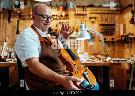 Side view of mature male luthier in apron and glasses sitting on chair and holding restored violin while working in workshop Stock Photo