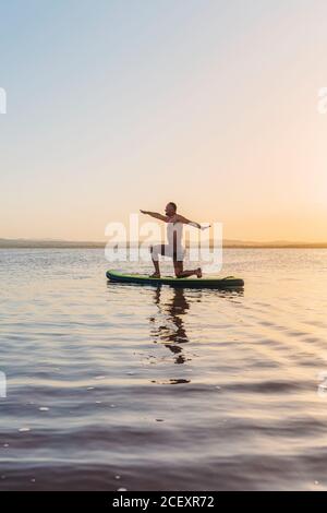 Full body side view of unrecognizable man doing variation of Crescent Lunge on Knee asana while floating on paddle board in calm lake water during sunset Stock Photo