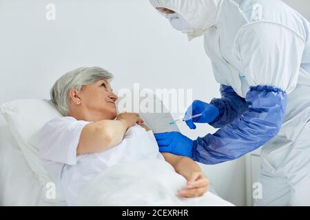 Modern doctor wearing protective suit giving injection to mature woman lying on bed in hospital ward Stock Photo