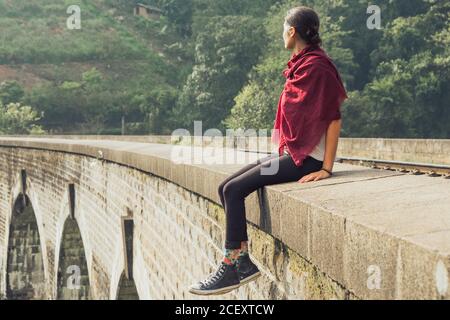 Low angle side view of thoughtful young ethnic female traveler in active wear and headscarf sitting on stone border of aged bridge against green tropical foliage during vacation in Sri Lanka Stock Photo
