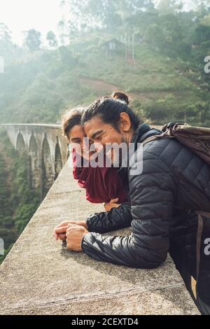 Side view of cheerful ethnic man and Woman in activewear with backpacks talking and laughing while standing on aged stone Nine Arch Bridge among green hills in Sri Lanka Stock Photo