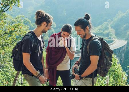 Group of cheerful young multiethnic friends with backpacks and photo camera having fun and laughing while standing on green hill near old viaduct bridge during summer holidays in Sri Lanka Stock Photo
