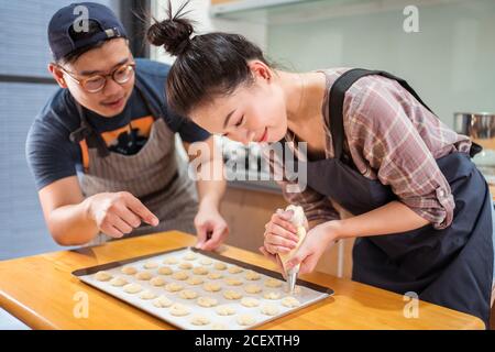 Asian guy helping smiling girlfriend with piping bag to pipe batter for pastries on baking pan at counter Stock Photo