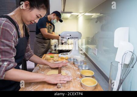 Side view of content Asian Woman in aprons rolling dough with rolling pin working at home near male cook on the background Stock Photo