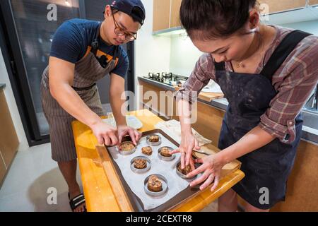 Crop Asian man and Woman spreading chocolate chip dough in tart rings on baking pan Stock Photo