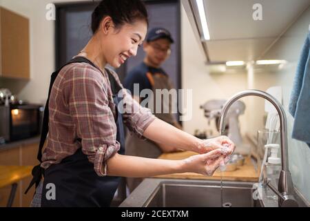 Side view of cheerful Asian Woman washing hands in sink while having home bakery with man Stock Photo