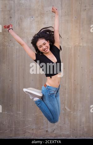 Side view of delighted Asian female celebrating victory and jumping with raised arms while looking at camera Stock Photo