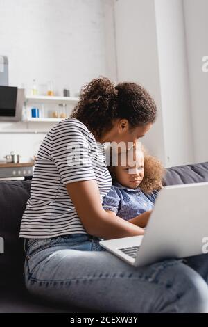 selective focus of african american freelancer kissing upset daughter while sitting on sofa with laptop Stock Photo