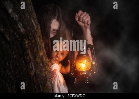 Scary woman with a lantern in night scene - Spooky image of a scary woman with dark eyes and appearance of a witch, in a white dress, holding a lit lantern and a frightening doll, in a dark night atmosphere .  Stock Photo
