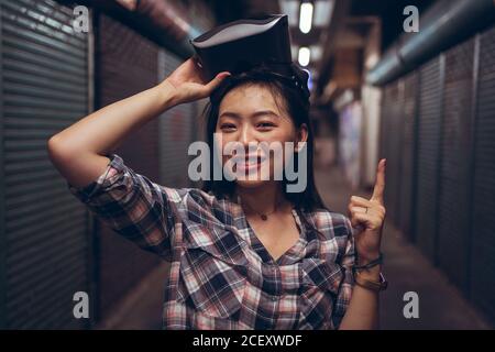Ethnic female taking off glasses of virtual reality and pointing up with index finger while looking at camera Stock Photo