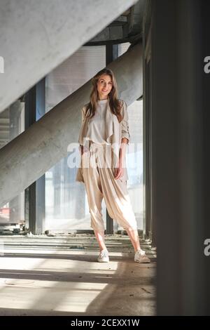 Full body of long haired blonde female in stylish blouse and pants looking at camera with smile while standing near contemporary concrete building Stock Photo