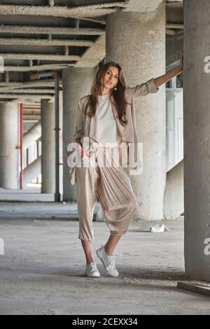 Full body of long haired blonde female in stylish blouse and pants looking at camera with smile while standing near contemporary concrete building Stock Photo