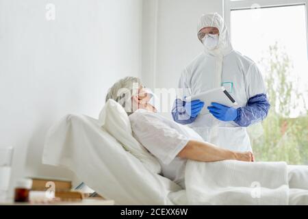 Unrecognizable man wearing protective suit with gloves, mask and eyewear holding digital tablet working with patient in infectious disease ward Stock Photo