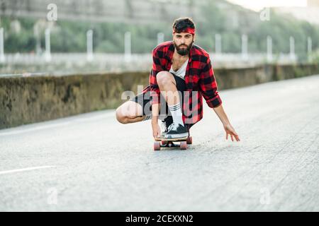 Male hipster in plaid shirt sitting on longboard and riding along road Stock Photo