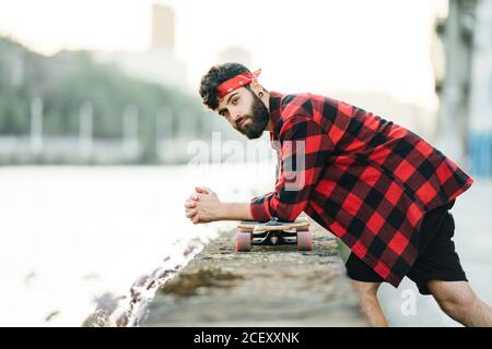 Side view of male hipster in checkered shirt leaning on longboard on stone border of embankment and looking at camera