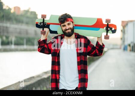 Cheerful bearded male hipster in bandana and checkered shirt standing on promenade with longboard and smiling at camera Stock Photo