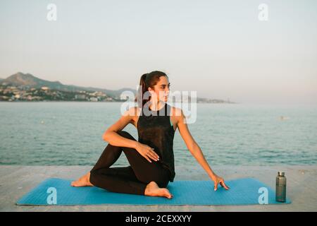 Young carefree female sitting on yoga mat in Ardha Matsyendrasana and stretching body while looking away on background of seascape in evening Stock Photo
