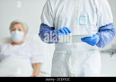 Horizontal medium shot of modern male nurse wearing protective uniform with gloves holding digital tablet with patient relaxing behind him Stock Photo