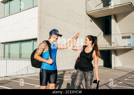 Smiling couple of athletes in sportswear standing on street and giving high five after training while looking at each other Stock Photo
