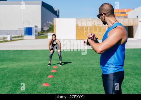 Side view of muscular male trainer noting time on wristwatch while female athlete preparing for running on green field with training cones Stock Photo