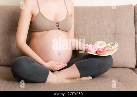 Close up of pregnant woman is eating many donuts relaxing on the sofa. Unhealthy dieting during pregnancy concept. Stock Photo