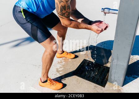 Side view of muscular tattooed athletic man in sportswear drinking refreshing cold water from street pump after hard training in city Stock Photo