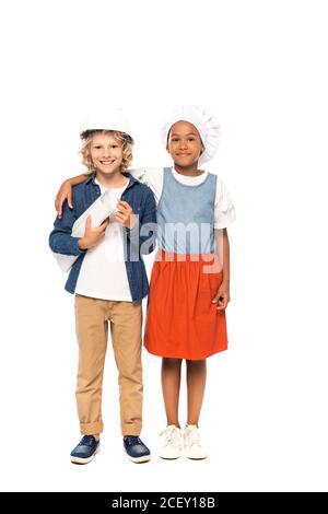 african american kid in chef hat hugging curly boy in safety helmet and costume of architect standing with blueprint isolated on white Stock Photo