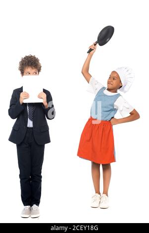 african american kid pretending housewife and holding frying pan near boy in suit covering face with digital tablet isolated on white Stock Photo