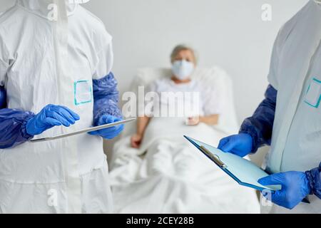 Two modern unrecognizable doctors wearing protective clothes standing in hospital ward with senior female patient on bed discussing something Stock Photo