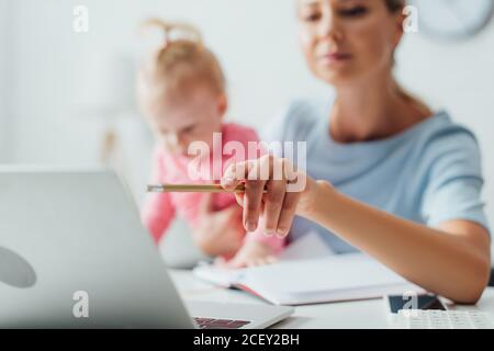 Selective focus of woman pointing with pencil at laptop while holding infant girl at home Stock Photo